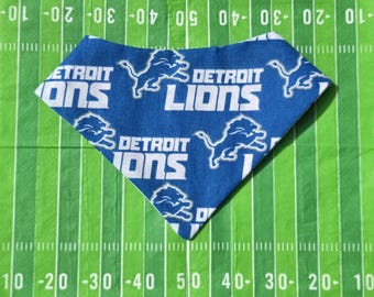 May include: A blue and white Detroit Lions pet bandana. The bandana features the team's logo and the words "Detroit Lions" in white. The background is a green football field.