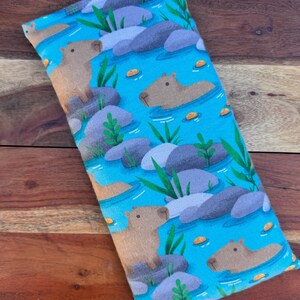 May include: A rectangular, light blue fabric item with a repeating pattern of capybaras in a river scene. The design includes gray rocks, green plants, and small orange floating objects. The capybaras are brown and tan.