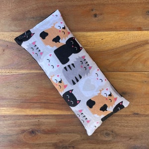 May include: White fabric eye pillow with a repeating pattern of black, gray, and brown cats with pink noses. The cats are all smiling and have different expressions.