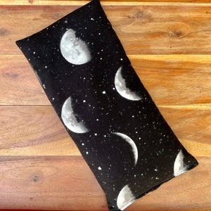 May include: A rectangular fabric pillow featuring a black background with a white celestial pattern of various moon phases and scattered stars. The textile has a soft texture and is oriented diagonally across the frame, displaying multiple lunar cycles.