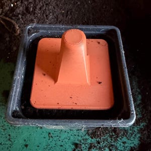 May include: An orange plastic soil tamper tool in a black plastic tray with soil. The tamper has a square base and a conical handle. It's used for compacting soil in gardening and is a useful tool.