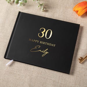 Op de afbeelding: Een zwart rechthoekig fotoalbum met goudkleurige tekst op de voorkant. De tekst luidt 30, HAPPY BIRTHDAY en de naam Emily in een cursief lettertype. Een klein wit lintje als bladwijzer steekt uit de onderkant van de zwarte kaft.