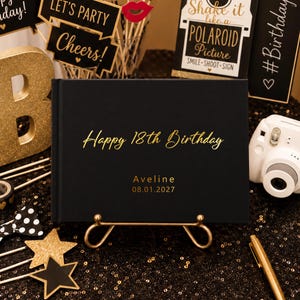 May include: A black rectangular guest book resting on a gold metal stand. The cover features gold foil text that reads Happy 18th Birthday, the name Aveline, and the date 08.01.2027. The book has a smooth matte finish and a hardbound spine.