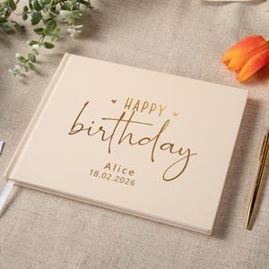 May include: A rectangular, cream-colored hardcover book with gold foil lettering on the front cover. The text reads "HAPPY birthday" with the name "Alice" and the date "18.02.2026" printed below in a clean, sans-serif font.
