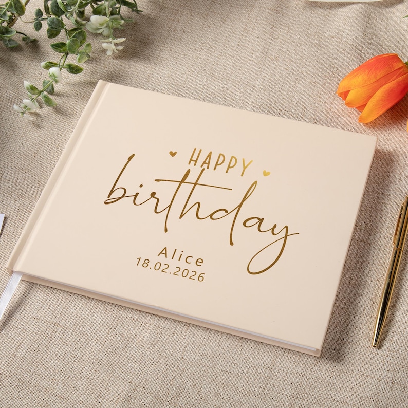 May include: A rectangular, cream-colored hardcover book with gold foil lettering on the front cover. The text reads "HAPPY birthday" with the name "Alice" and the date "18.02.2026" printed below in a clean, sans-serif font.