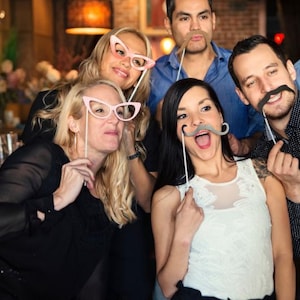 May include: A collection of photo booth props including pink cat-eye glasses, a black mustache, a brown mustache, and a gray mustache, each attached to a thin white stick, held up by a group of people posing for a photograph in a dimly lit setting.
