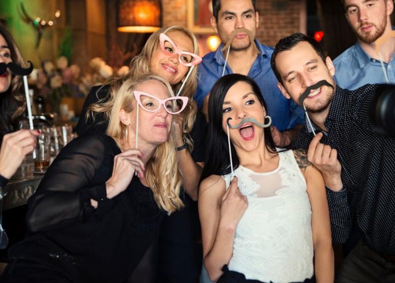 May include: A collection of photo booth props including pink cat-eye glasses, a black mustache, a brown mustache, and a gray mustache, each attached to a thin white stick, held up by a group of people posing for a photograph in a dimly lit setting.