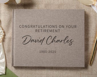 May include: A rectangular, light brown, fabric-covered guest book featuring the dark text "CONGRATULATIONS ON YOUR RETIREMENT", "David Charles", and the years "1985-2025" printed in a combination of sans-serif and script fonts on the front cover.