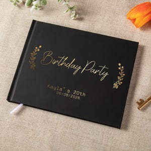 May include: A black rectangular photo album with gold foil text that reads Birthday Party, Kayla's 20th, and 06/08/2026. The cover features gold botanical illustrations on both sides of the title and includes a small white ribbon bookmark.