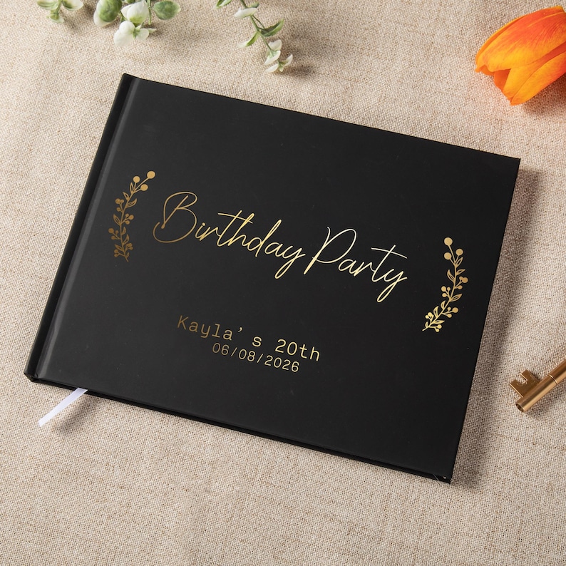 May include: A black rectangular photo album with gold foil text that reads Birthday Party, Kayla's 20th, and 06/08/2026. The cover features gold botanical illustrations on both sides of the title and includes a small white ribbon bookmark.