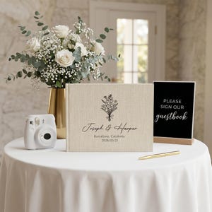 Puede incluir: Un libro de visitas de boda beige con los nombres "Joseph & Harper" y la fecha "2026/03/21" est&aacute; sobre un mantel blanco. Tambi&eacute;n hay una c&aacute;mara instant&aacute;nea blanca, un jarr&oacute;n dorado con rosas blancas y un letrero que dice "Please sign our guestbook".