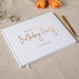May include: A white rectangular guest book with gold foil lettering that reads ANDRE'S 21ST Birthday Party and the date 06/09/2026. A gold pen is placed on the right side of the book cover, which features a smooth finish and a white ribbon bookmark.