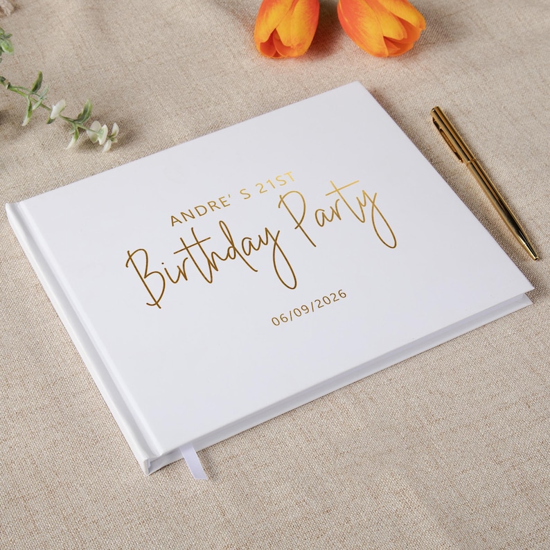 May include: A white rectangular guest book with gold foil lettering that reads ANDRE'S 21ST Birthday Party and the date 06/09/2026. A gold pen is placed on the right side of the book cover, which features a smooth finish and a white ribbon bookmark.