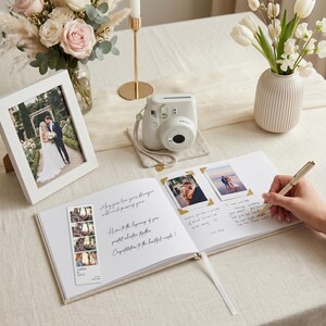 Puede incluir: Un libro de visitas de boda, una c&aacute;mara instant&aacute;nea blanca y una foto enmarcada est&aacute;n dispuestas sobre una mesa. El libro est&aacute; abierto, con fotos y mensajes manuscritos. Un bol&iacute;grafo est&aacute; sobre la p&aacute;gina. Flores y una vela completan la escena.