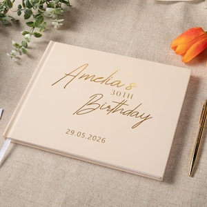 May include: A light beige rectangular guest book with gold foil lettering that reads Amelia's 30TH Birthday and the date 29.05.2026. The cover features a smooth finish and is positioned next to a gold pen and a small orange flower petal.