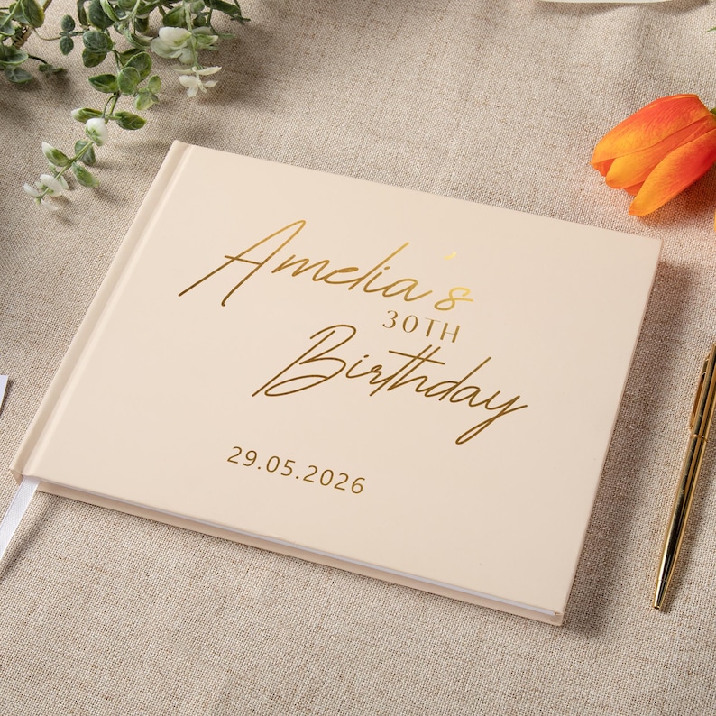 May include: A light beige rectangular guest book with gold foil lettering that reads Amelia's 30TH Birthday and the date 29.05.2026. The cover features a smooth finish and is positioned next to a gold pen and a small orange flower petal.