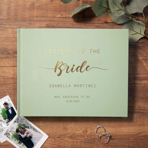 Può includere: Un libro degli ospiti di nozze verde chiaro con scritte dorate. È personalizzato con il nome della sposa, il suo futuro nome da sposata e la data del matrimonio. Sono visibili anche due fedi nuziali e due foto di matrimonio.