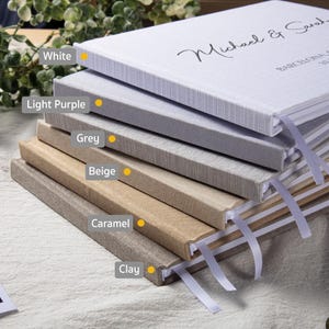 Puede incluir: Una pila de libros de visitas de tapa dura en varios colores neutros: blanco, lila claro, gris, beige, caramelo y arcilla. Cada libro tiene un marcador de cinta. El libro superior est&aacute; inscrito con los nombres "Michael & Sarah" y la ciudad "Barcelona".