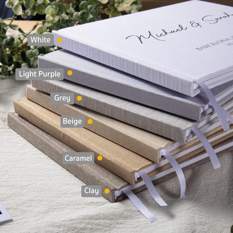 Puede incluir: Una pila de libros de visitas de tapa dura en varios colores neutros: blanco, lila claro, gris, beige, caramelo y arcilla. Cada libro tiene un marcador de cinta. El libro superior est&aacute; inscrito con los nombres "Michael & Sarah" y la ciudad "Barcelona".