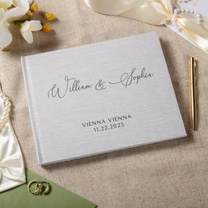 Puede incluir: Un libro de visitas de boda de color gris claro con textura de lino y los nombres "William & Sophia" en escritura elegante. Debajo, las palabras "VIENNA VIENNA" y la fecha "11.22.2025". Un bol&iacute;grafo dorado descansa junto al libro.