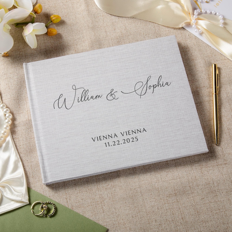 Puede incluir: Un libro de visitas de boda de color gris claro con textura de lino y los nombres "William & Sophia" en escritura elegante. Debajo, las palabras "VIENNA VIENNA" y la fecha "11.22.2025". Un bol&iacute;grafo dorado descansa junto al libro.
