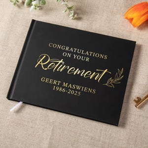 May include: A black rectangular guest book with gold foil lettering that reads CONGRATULATIONS ON YOUR RETIREMENT, GEERT MASWIENS, 1986-2025. The cover features a decorative gold leaf branch illustration and a small white ribbon bookmark.