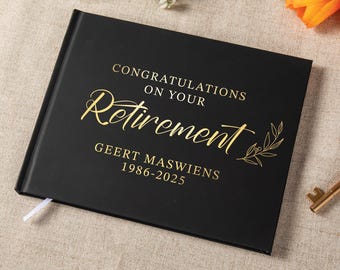 May include: A black rectangular guest book with gold foil lettering that reads CONGRATULATIONS ON YOUR RETIREMENT, GEERT MASWIENS, 1986-2025. The cover features a decorative gold leaf branch illustration and a small white ribbon bookmark.