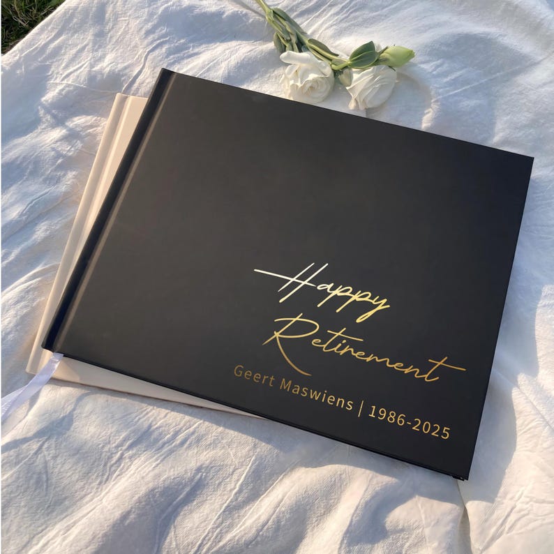 May include: A black rectangular photo album with gold foil lettering that reads Happy Retirement Geert Maswiens | 1986-2025. The album is positioned on top of a light beige album, with white flowers placed near the top edge of the black cover.