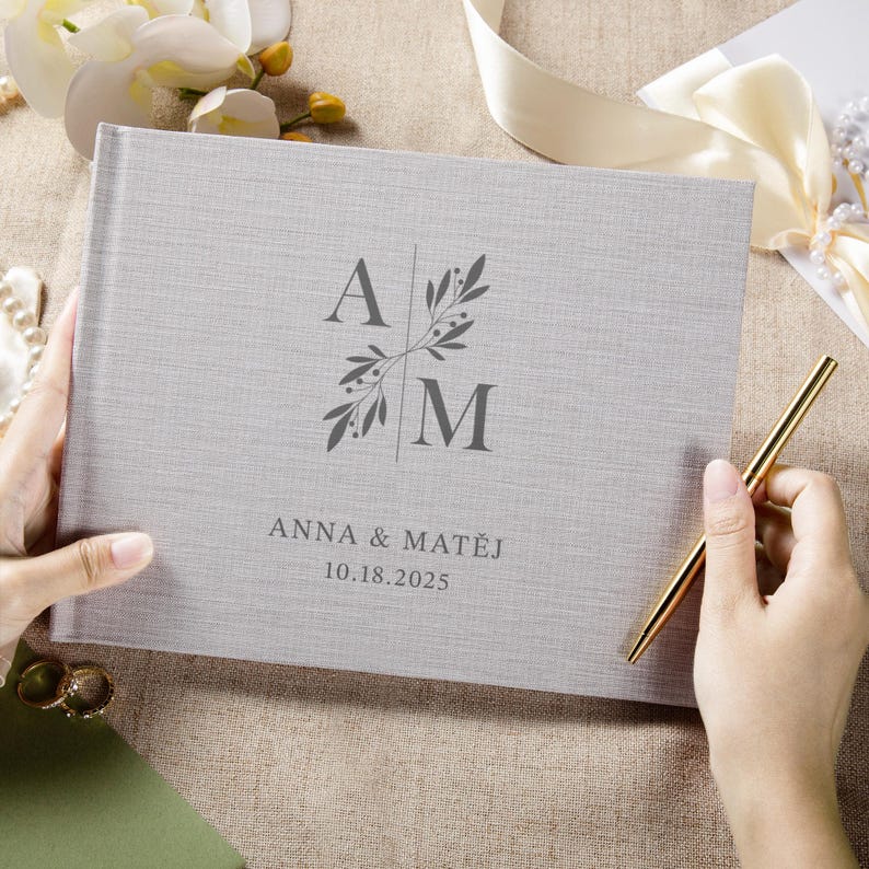 Puede incluir: Un libro de visitas de boda de color gris claro con textura de lino, con las iniciales "A" y "M" y los nombres "Anna & Matej" y la fecha "10.18.2025" impresos en la portada. Un bol&iacute;grafo dorado descansa sobre el libro.