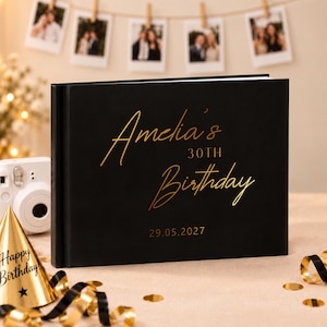 May include: A black photo album with gold foil text that reads Amelia's 30TH Birthday and the date 29.05.2027. The rectangular book features a smooth matte cover and is positioned next to a white instant camera and a gold party hat with black ribbon.