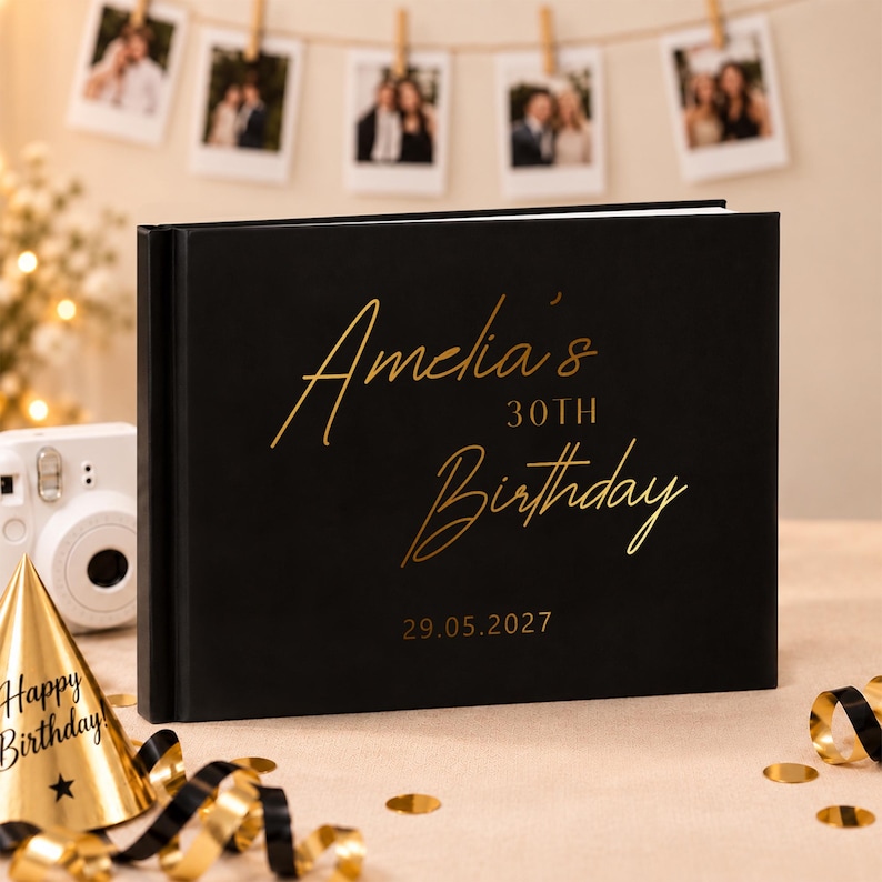 May include: A black photo album with gold foil text that reads Amelia's 30TH Birthday and the date 29.05.2027. The rectangular book features a smooth matte cover and is positioned next to a white instant camera and a gold party hat with black ribbon.