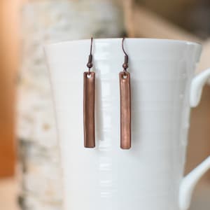 May include: A pair of copper-colored dangle earrings with a rectangular shape. The earrings are hanging from a white mug.