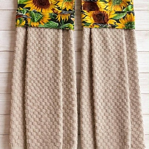 May include: Two beige kitchen towels with a textured pattern. The top of each towel features a sunflower and bird print on a black background. The text "Sunflower & birds 18" is at the bottom.