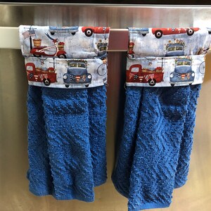 May include: Two blue kitchen towels with red, white and blue patterned fabric tops. The fabric features a vintage truck design with the words "Welcome Home" on one of the trucks. The towels are hanging on a silver bar.