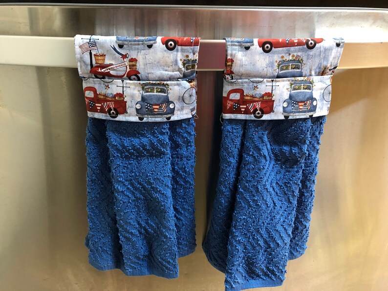 May include: Two blue kitchen towels with red, white and blue patterned fabric tops. The fabric features a vintage truck design with the words "Welcome Home" on one of the trucks. The towels are hanging on a silver bar.