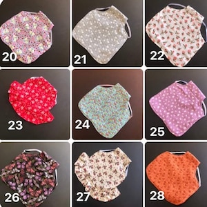 May include: A collection of nine fabric bibs labeled 20 through 28, each featuring distinct patterns including butterflies, floral prints, and solid colors. These bibs are shaped with rounded edges and include elastic neck straps for secure fastening.
