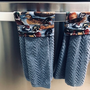 May include: Two blue kitchen towels with a duck print border hanging on a silver towel rack. The towels are made of a soft, absorbent material.