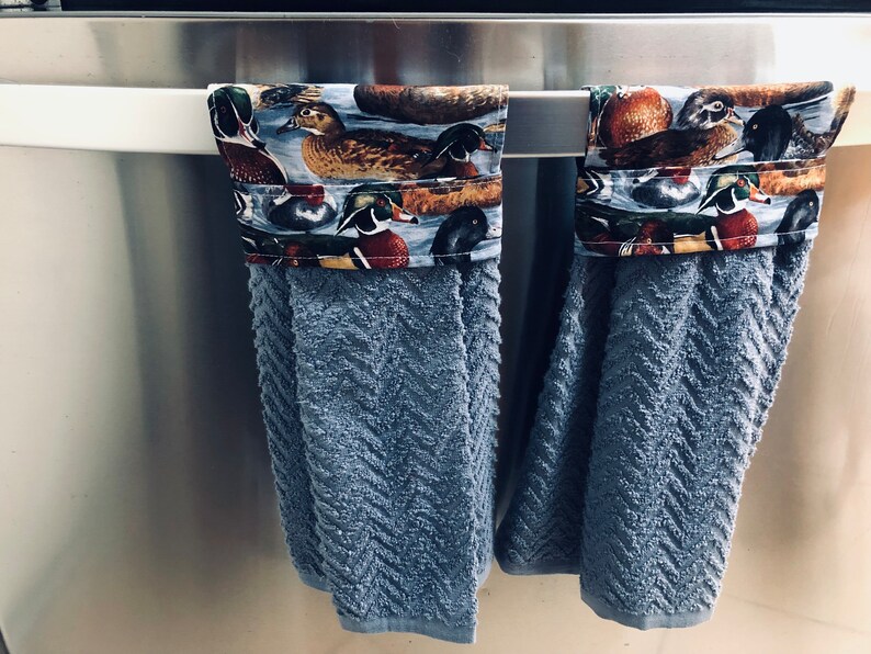 May include: Two blue kitchen towels with a duck print border hanging on a silver towel rack. The towels are made of a soft, absorbent material.