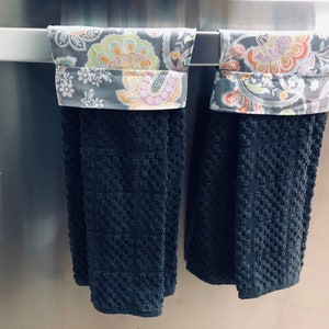 May include: Two black kitchen towels with a floral patterned fabric trim. The towels are hanging on a silver towel rack.