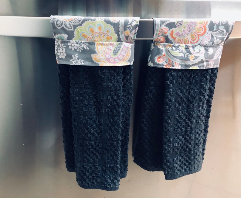 May include: Two black kitchen towels with a floral patterned fabric trim. The towels are hanging on a silver towel rack.
