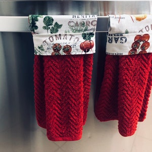 May include: Two red kitchen towels with white fabric trim featuring a garden-themed print with the words "Tomato", "Garlic", and "Caprico" hanging on a silver bar.