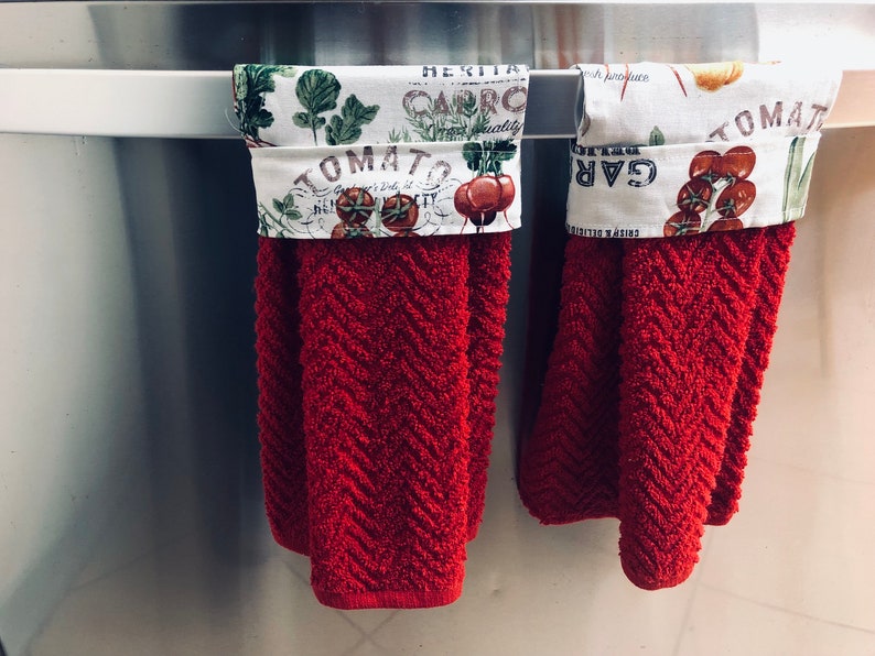 May include: Two red kitchen towels with white fabric trim featuring a garden-themed print with the words "Tomato", "Garlic", and "Caprico" hanging on a silver bar.