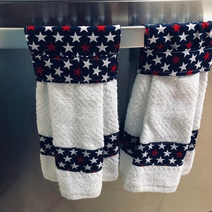 May include: Two white kitchen towels with red and blue star patterned fabric accents. The towels are hanging on a silver towel rack.