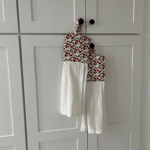 May include: Two white kitchen towels with a red cherry print. The towels are hanging on a white cabinet door.