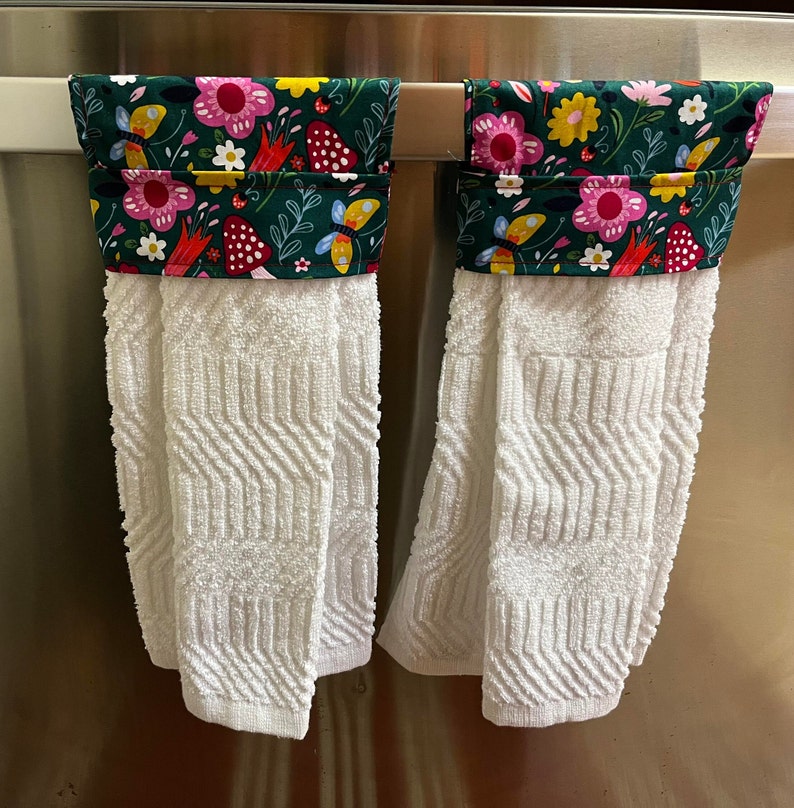 May include: Two white kitchen towels with green, pink, yellow, and red floral print fabric hanging on a silver towel rack.