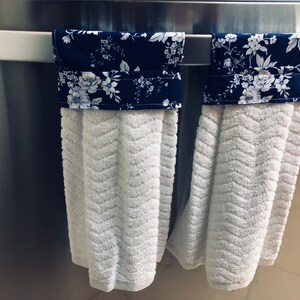 May include: Two white kitchen towels with blue and white floral fabric accents hanging on a silver towel rack.