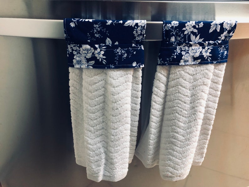 May include: Two white kitchen towels with blue and white floral fabric accents hanging on a silver towel rack.