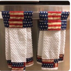 May include: Two white kitchen towels with red, white and blue patterned borders hanging on a silver towel rack. The towels have a textured weave.
