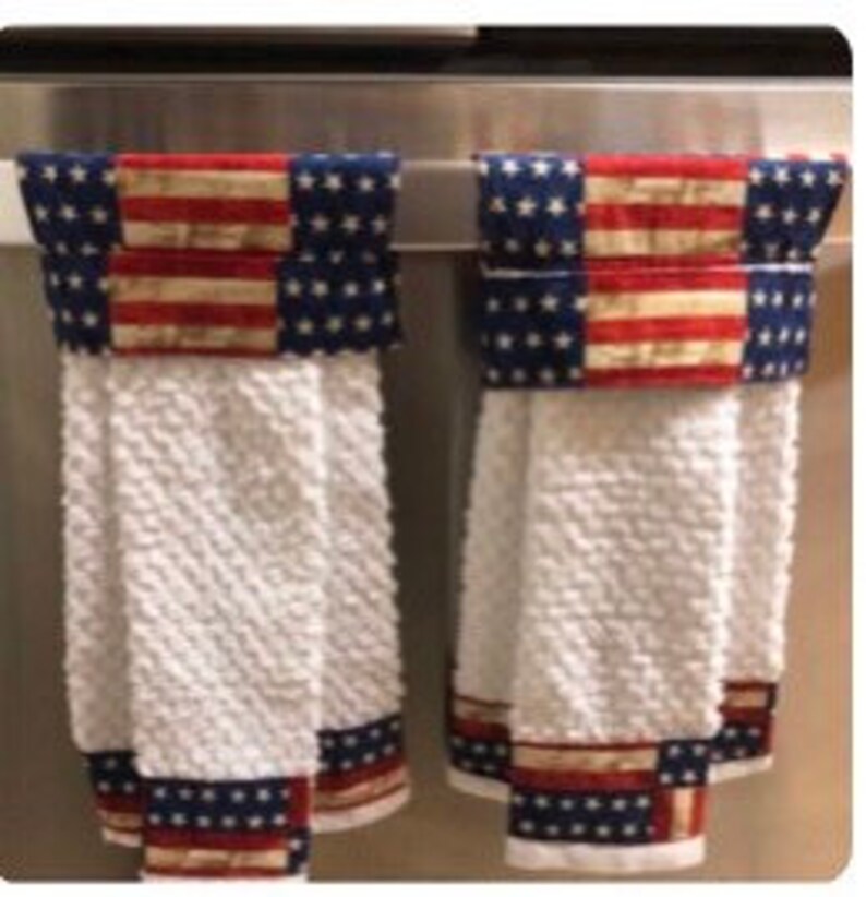 May include: Two white kitchen towels with red, white and blue patterned borders hanging on a silver towel rack. The towels have a textured weave.