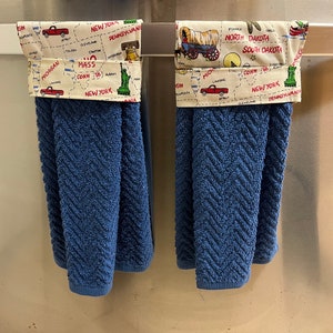 May include: Two blue kitchen towels with a white and yellow patterned fabric border. The border features a map of the United States with various state names and landmarks. One towel has a red truck and the other has a covered wagon.